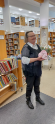 Kuvassa Vuoden lainaajaksi valittu henkilö, jolla on kukkia ja suklaarasia käsissään. Kuva on otettu kirjaston sisätiloissa ja henkilön ympärillä on kirjahyllyjä sekä kirjakärryjä.